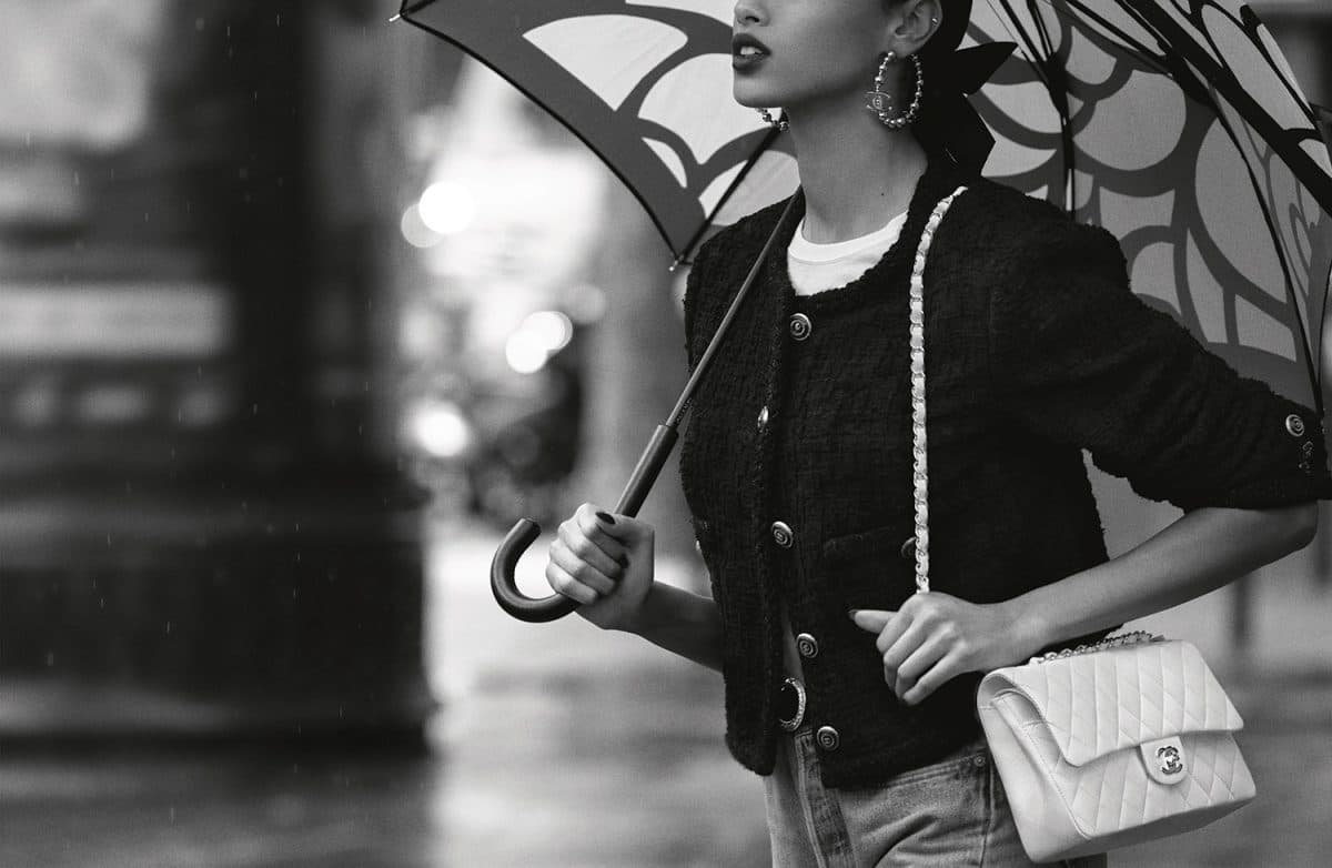 Model Imaan Hammam holding an umbrella and wearing a classic Chanel tweed jacket with a white quilted Chanel Flap Bag.